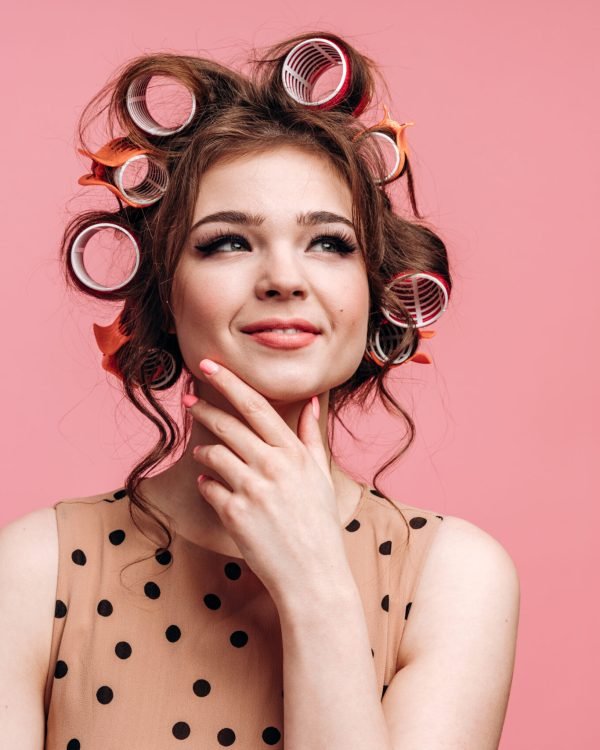 Beautiful brunette in hair curlers gently touches her face on a pink background. Isolated on empty wall background.